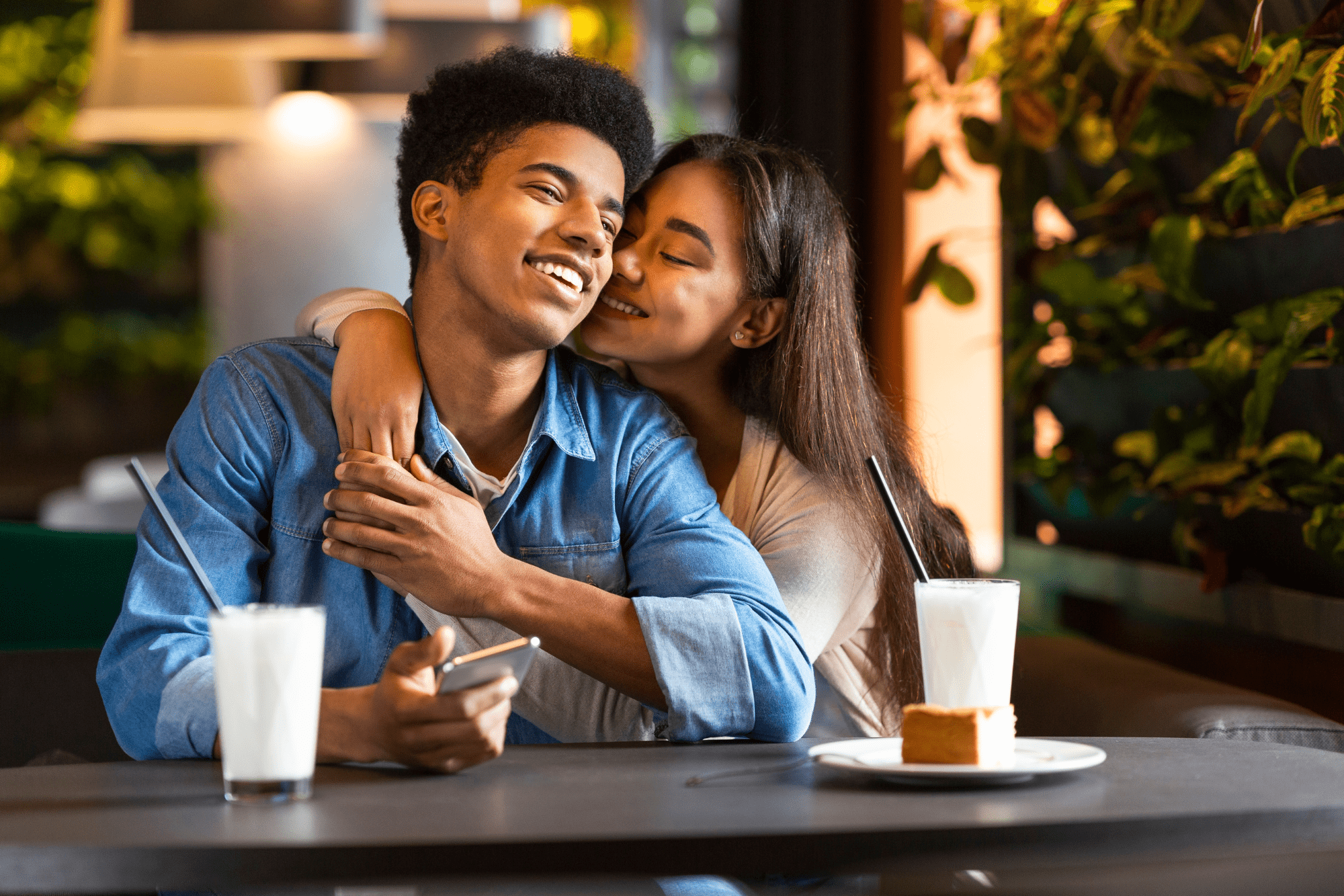 A black man and an asian woman sit at a cafe table. She is leaning affectionately on his shoulder.