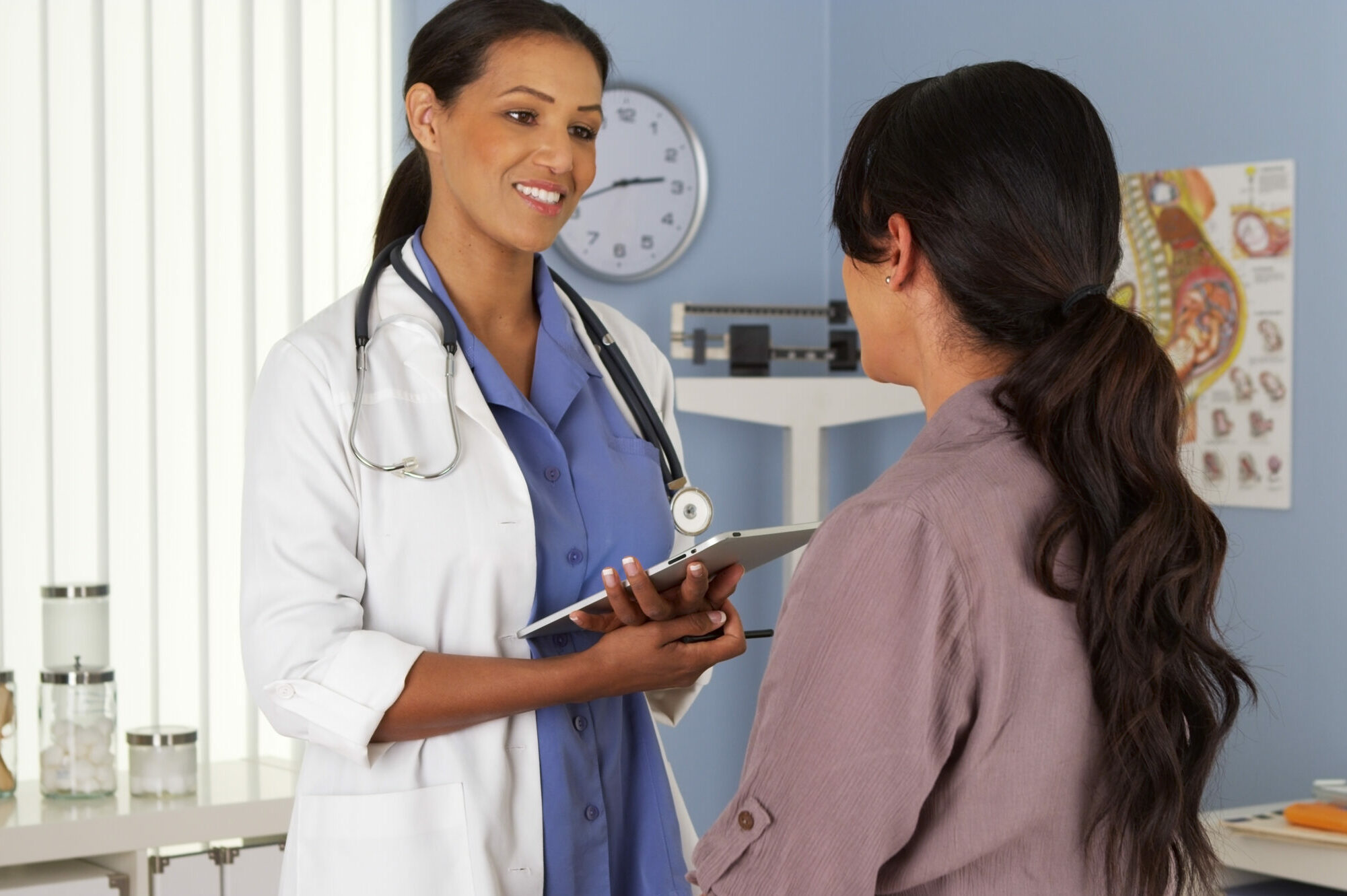 A black woman doctor speaking to a patient in a blue exam room.